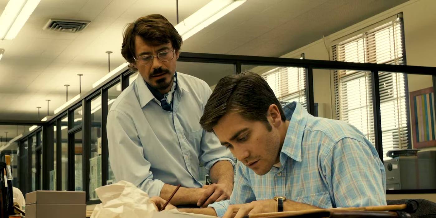 Thrillers psicológicos: 10 filmes com roteiro impecável Robert Graysmith (Jake Gyllenhaal) curvado sobre sua mesa enquanto Paul Avery (Robert Downey Jr.) fica casualmente atrás dele em 'Zodiac' (2007).